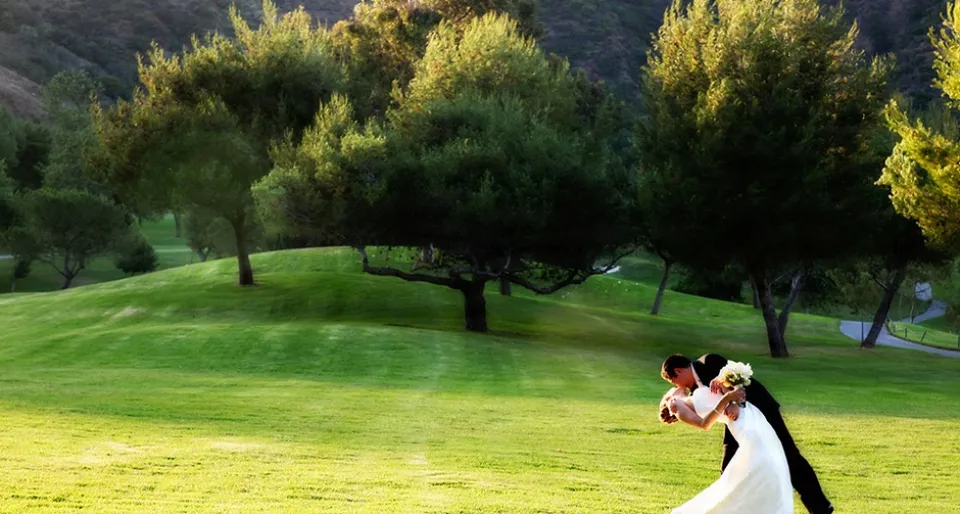 Groom dipping and kissing his bride on a green lawn with oak trees and hills behind in warm light.
